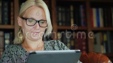 戴眼镜，坐在椅子上，用平板电脑的女人的肖像。 在书架的背景下
